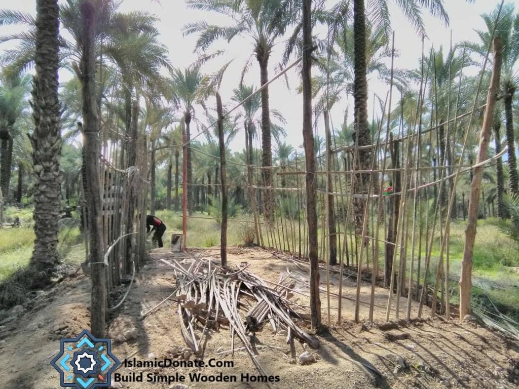 A laborer is building a simple wooden fence in a date palm grove, with piles of wood nearby. This project supports families with income and shelter through date tree cultivation and provides aid via RLUSD crypto donations.