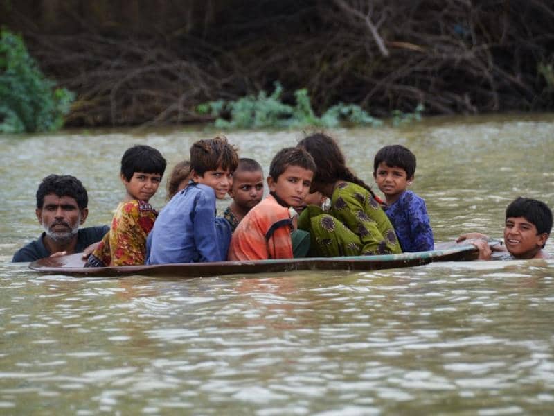 A man and several children crammed into a boat navigating floodwaters in Pakistan, with **USDC** donation aiding relief efforts.