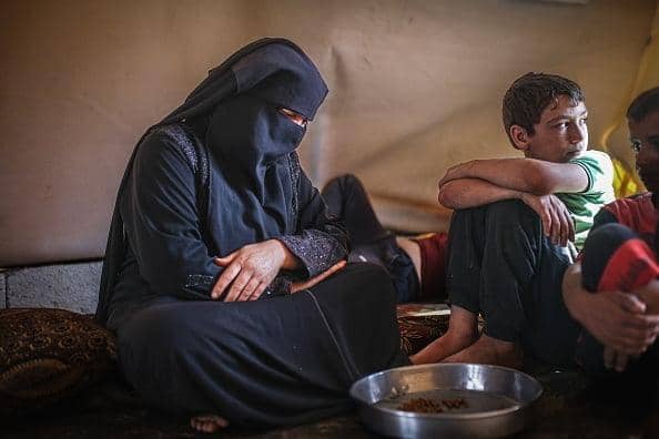 A veiled Syrian woman sits with her arms crossed, looking down, beside children in a makeshift shelter. Relief efforts supported by cryptocurrency donations, possibly using Ethereum, aim to rebuild lives.