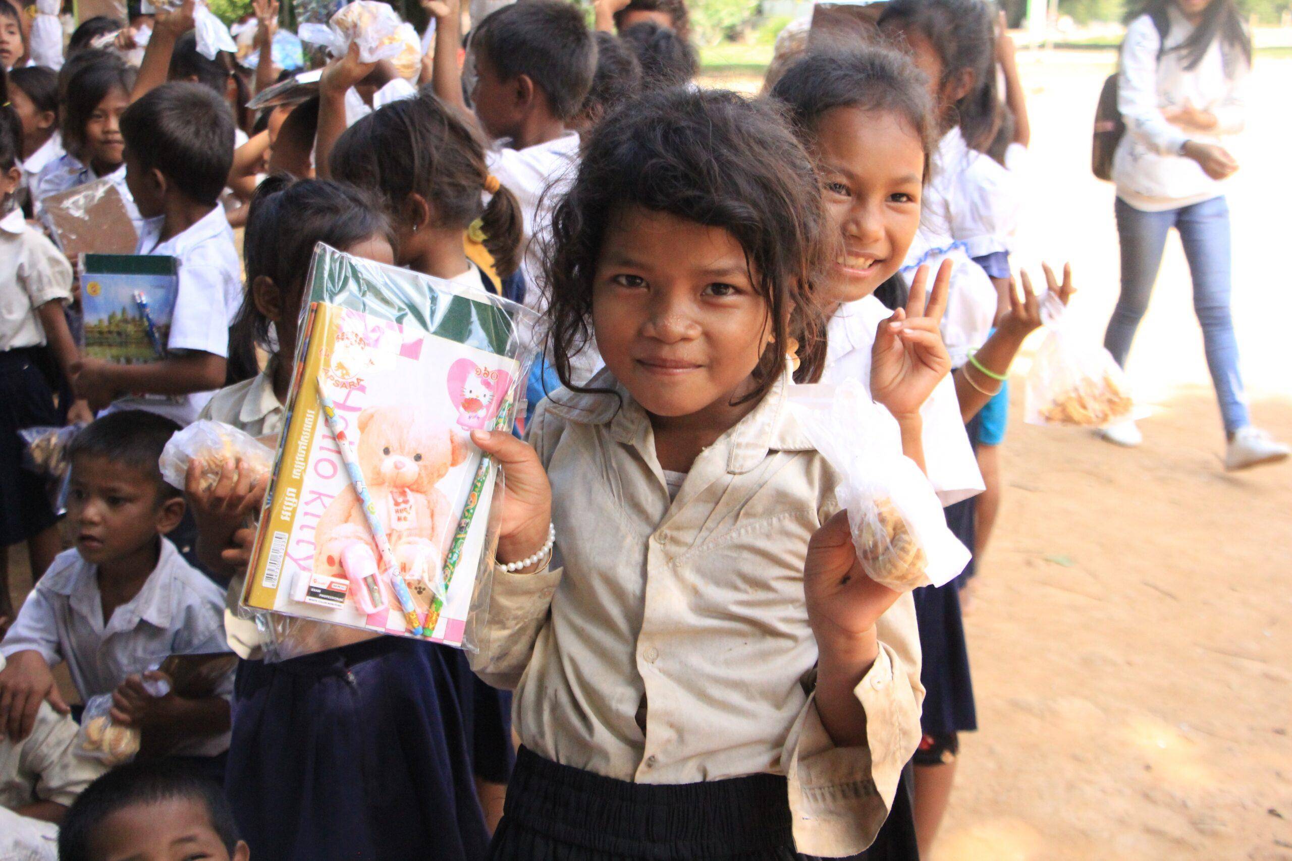 Children in relief line holding school supplies and snacks, with a child in the foreground smiling and holding a Hello Kitty binder, receiving aid via Litecoin.
