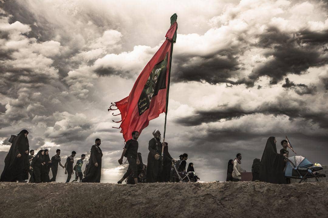 A pilgrimage procession walks under a dramatic sky, led by a large red flag. This scene depicts a journey of faith and community, perhaps supported by crypto donations via ETH.