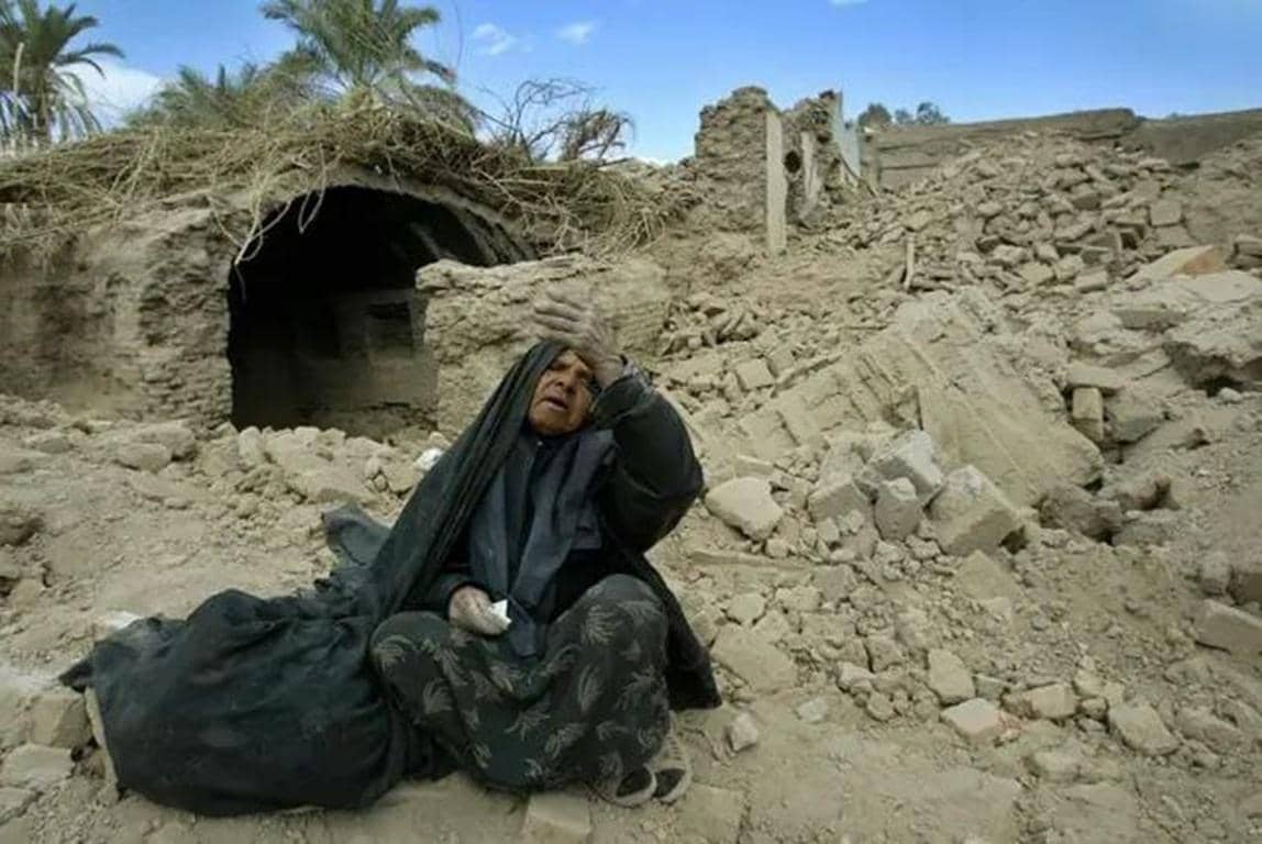Elderly woman in mourning attire sits amidst rubble with her hand to her forehead, a symbol of the plight addressed by crypto zakat donations for relief in Palestine and Yemen.