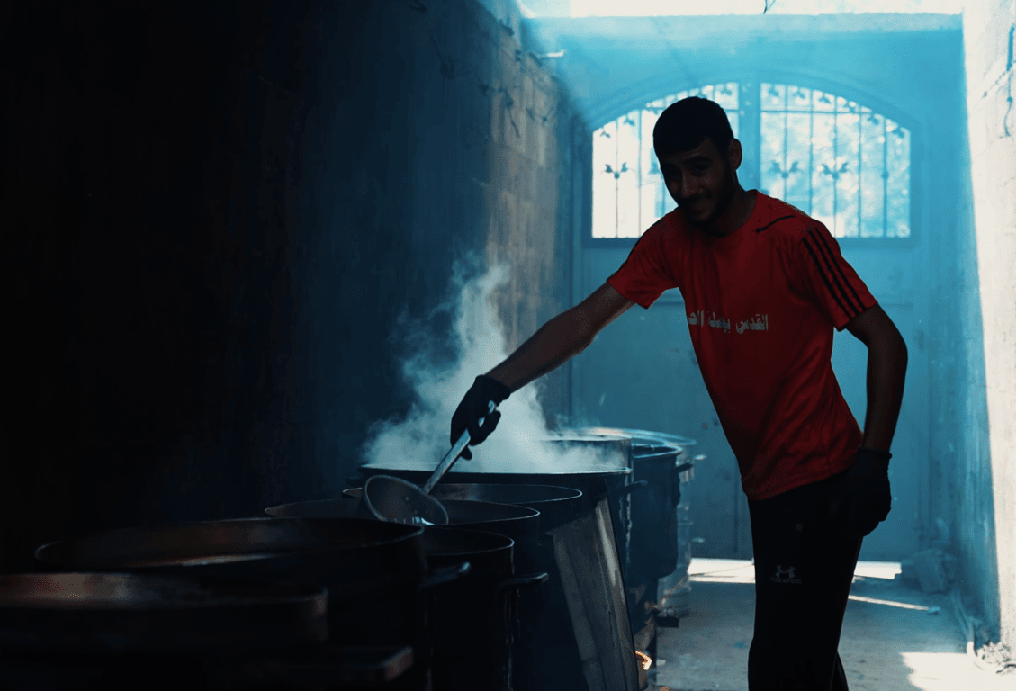 A young man in a red shirt stirs a large pot of food in a steamy kitchen, distributing aid for Palestine. Donate crypto with Dogecoin.