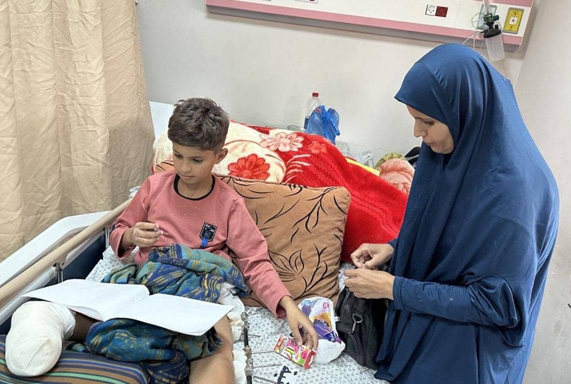 A volunteer in a blue hijab provides comfort and items to a young boy with a bandaged leg who is sitting up in a hospital bed, reading a book. Charitable aid for displaced Palestinian families, with opportunities to donate cryptocurrency like USDT or USDC.