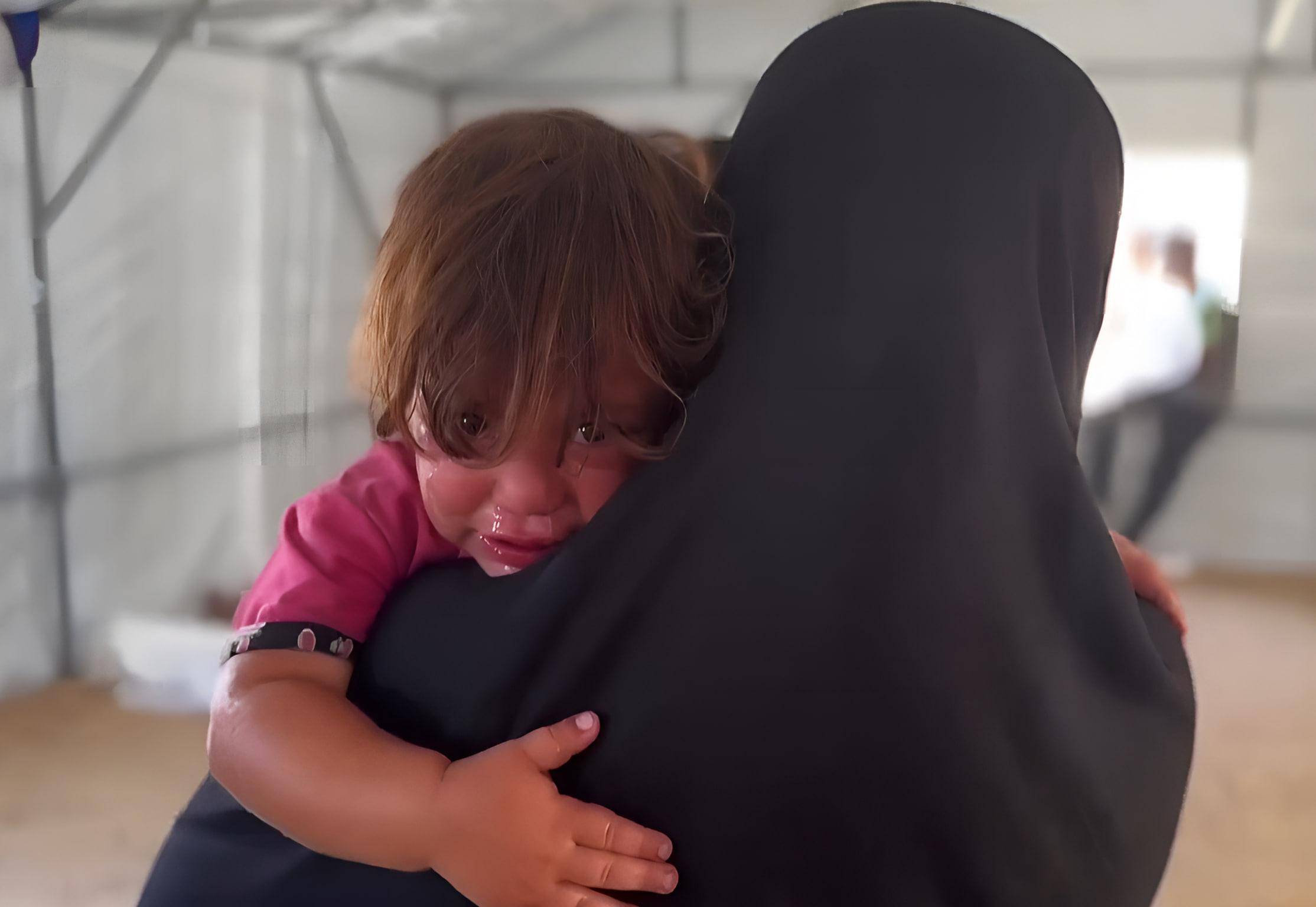 Close-up of a crying child being held by a mother wearing a black hijab. The child's face is wet with tears, and their hand rests on the mother's shoulder. Urgent need for aid in Palestine, with donations via BTC.