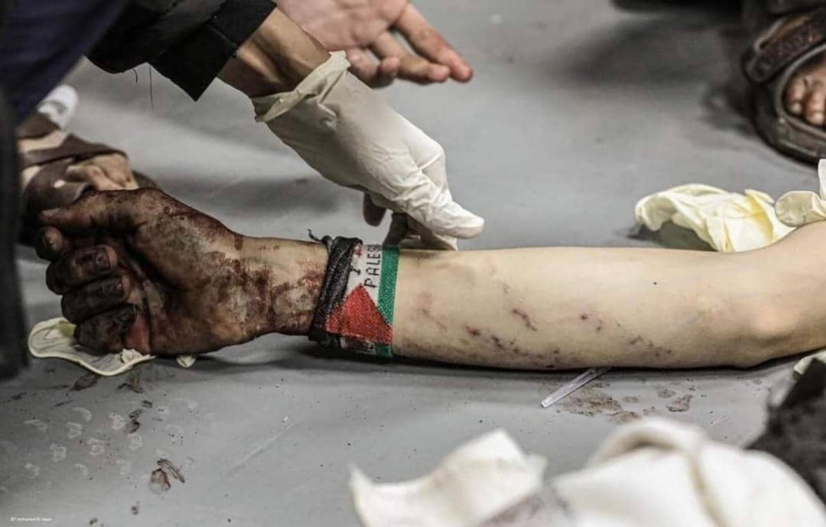 Medical professional's gloved hands attend to an injured arm, a Palestinian flag bracelet is visible. Donate crypto like Solana for urgent relief in Palestine.