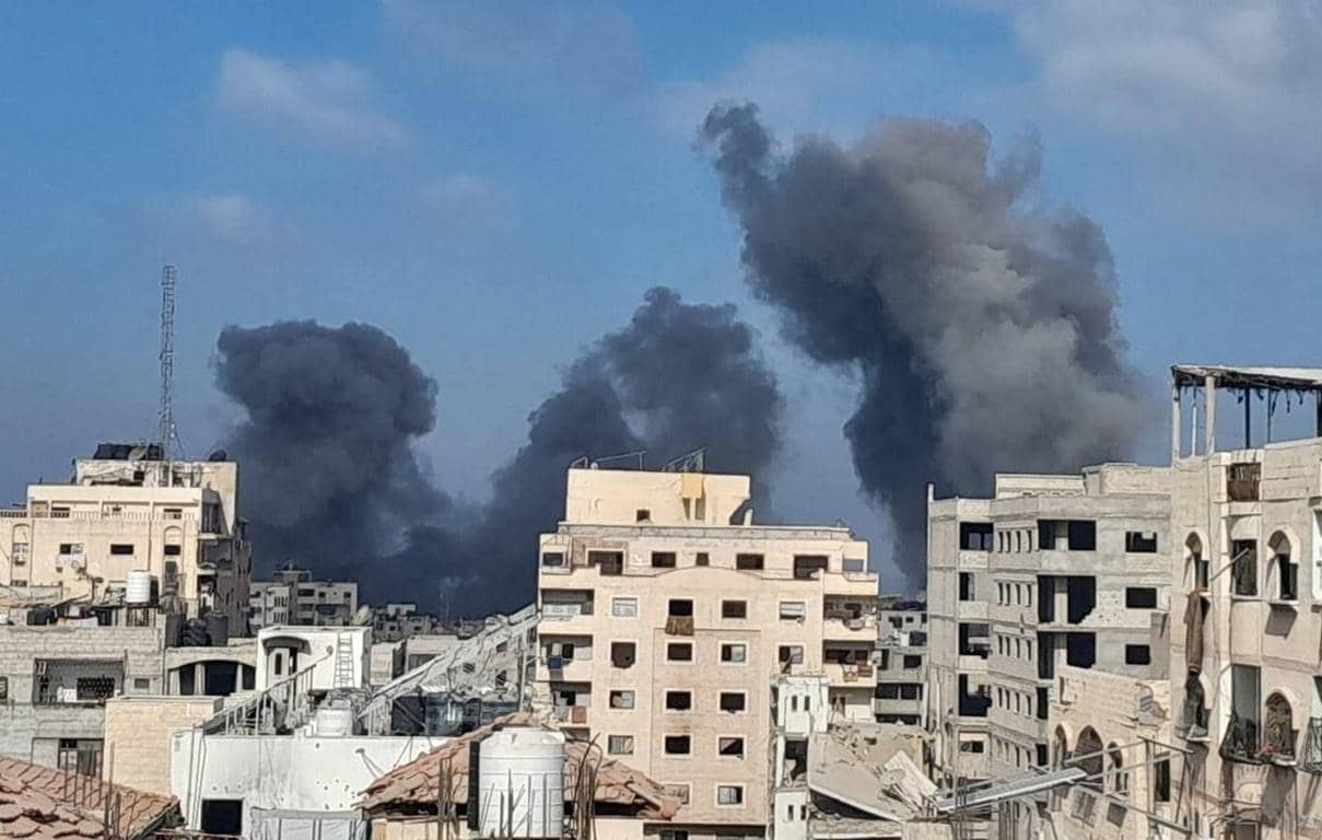 Thick plumes of black smoke rise above buildings in a city, indicating ongoing destruction and humanitarian crisis in Palestine. Cryptocurrencies like RLUSD can help fund urgent relief.