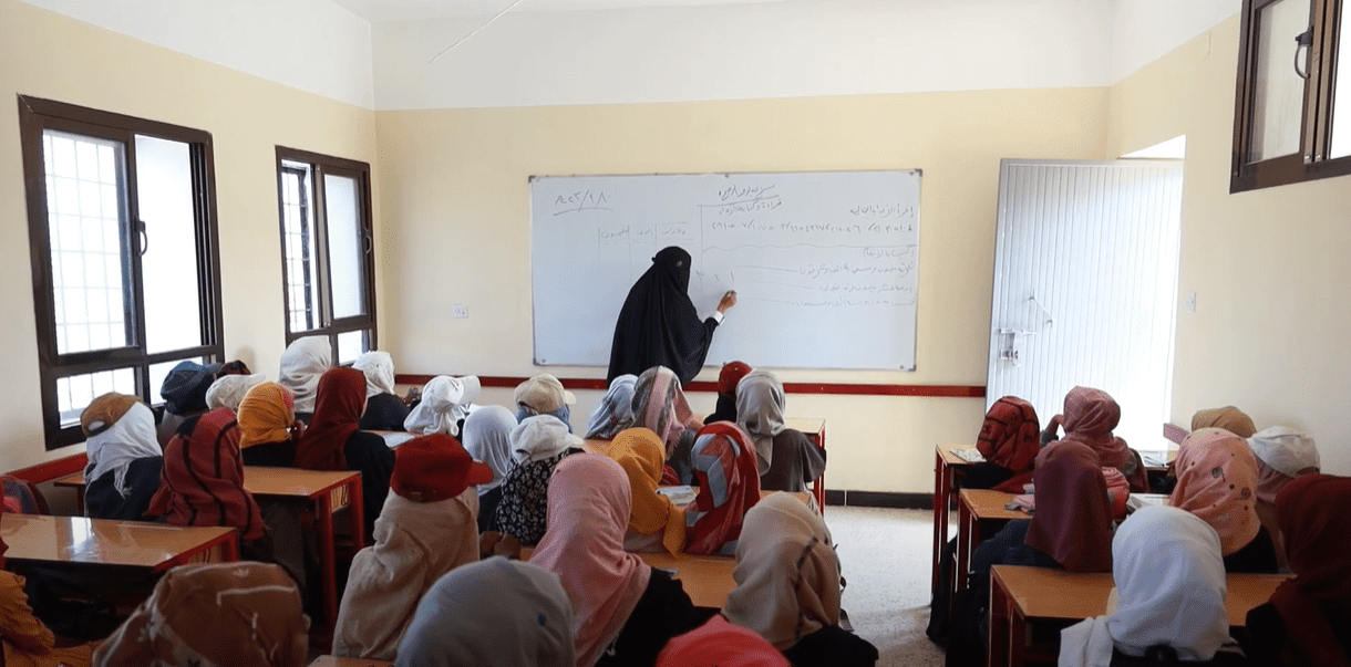 Classroom of young girls in headscarves learning from a teacher writing on a whiteboard, supported by crypto donations like ETH.