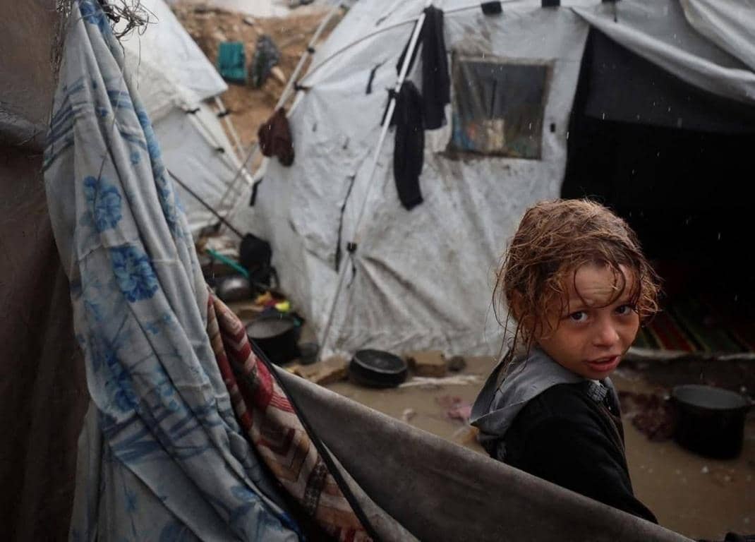 Child in a refugee camp with worn tents behind, looking at the camera. Winter relief aid sponsored by crypto donations in BTC.