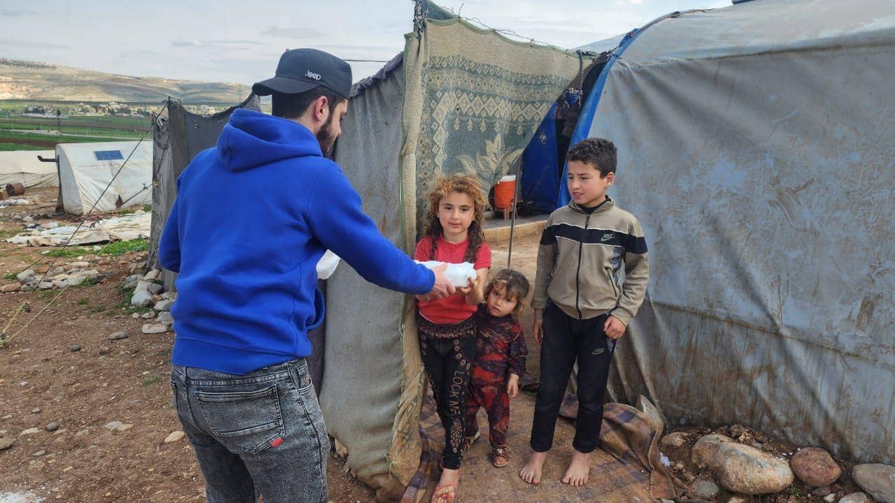 A volunteer distributes aid to children near tents in Aleppo, with support facilitated by USDT. Aid includes food, medicine, shelter, and water, embodying Islamic principles of charity. Keywords: Aleppo crisis, humanitarian aid, crypto donations, Islamic charity, poverty and hunger alleviation.