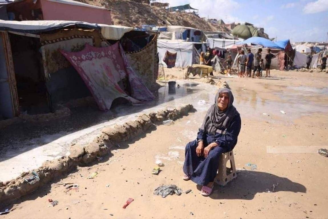 Elderly woman in a refugee camp, with makeshift shelters behind her. Your crypto zakat, for example, via ETH, can provide essential aid and hope to those affected by crises.