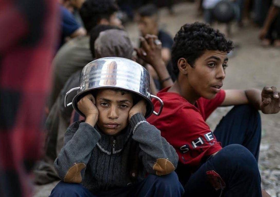 A child wearing a cooking pot on their head sits with hands on cheeks while another teen looks away, symbolizing need. Crypto Zakat donation supports relief efforts.
