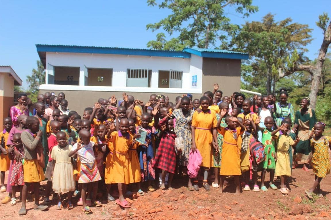Children waving cheerfully outside a newly built school in Uganda, a project benefiting from cryptocurrency donations for clean water and community empowerment.