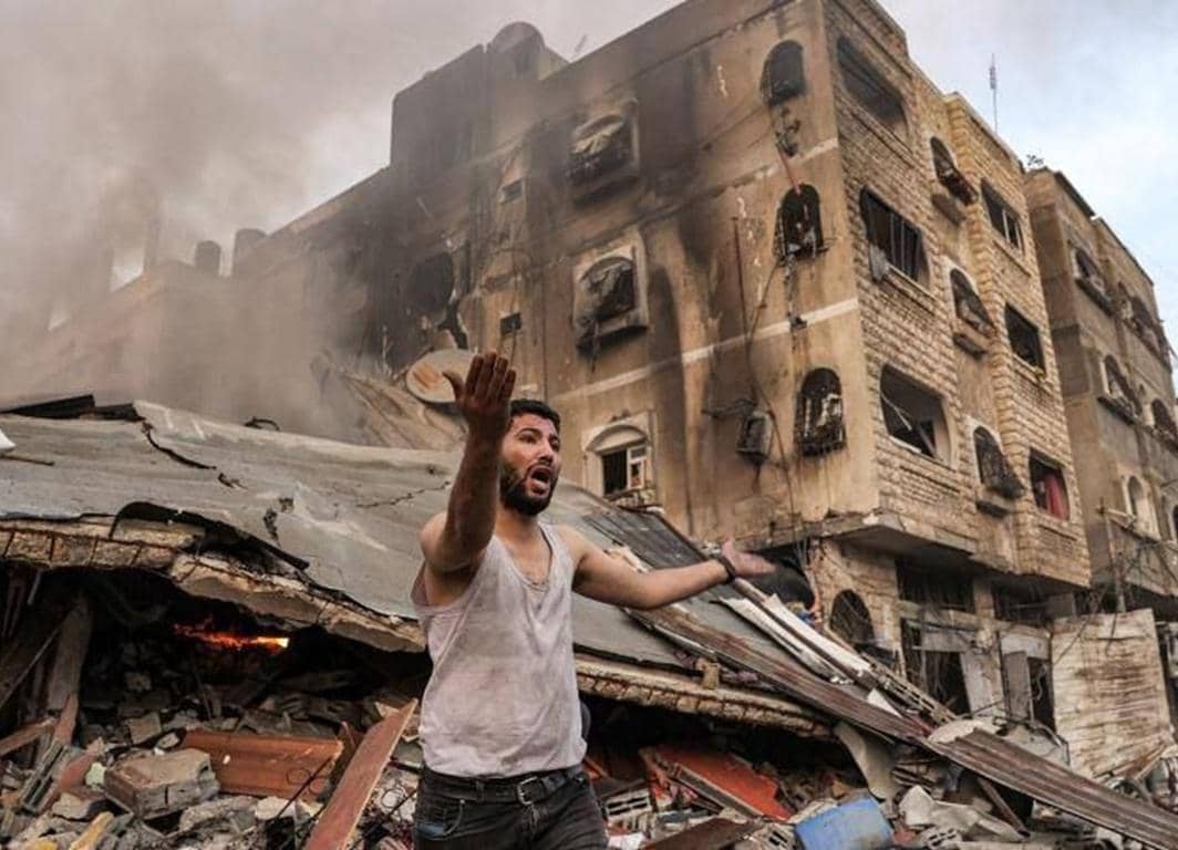 A distressed man gestures in front of a heavily damaged, smoke-billowing building in Palestine, highlighting the urgent need for crypto Zakat donations for reconstruction and aid.