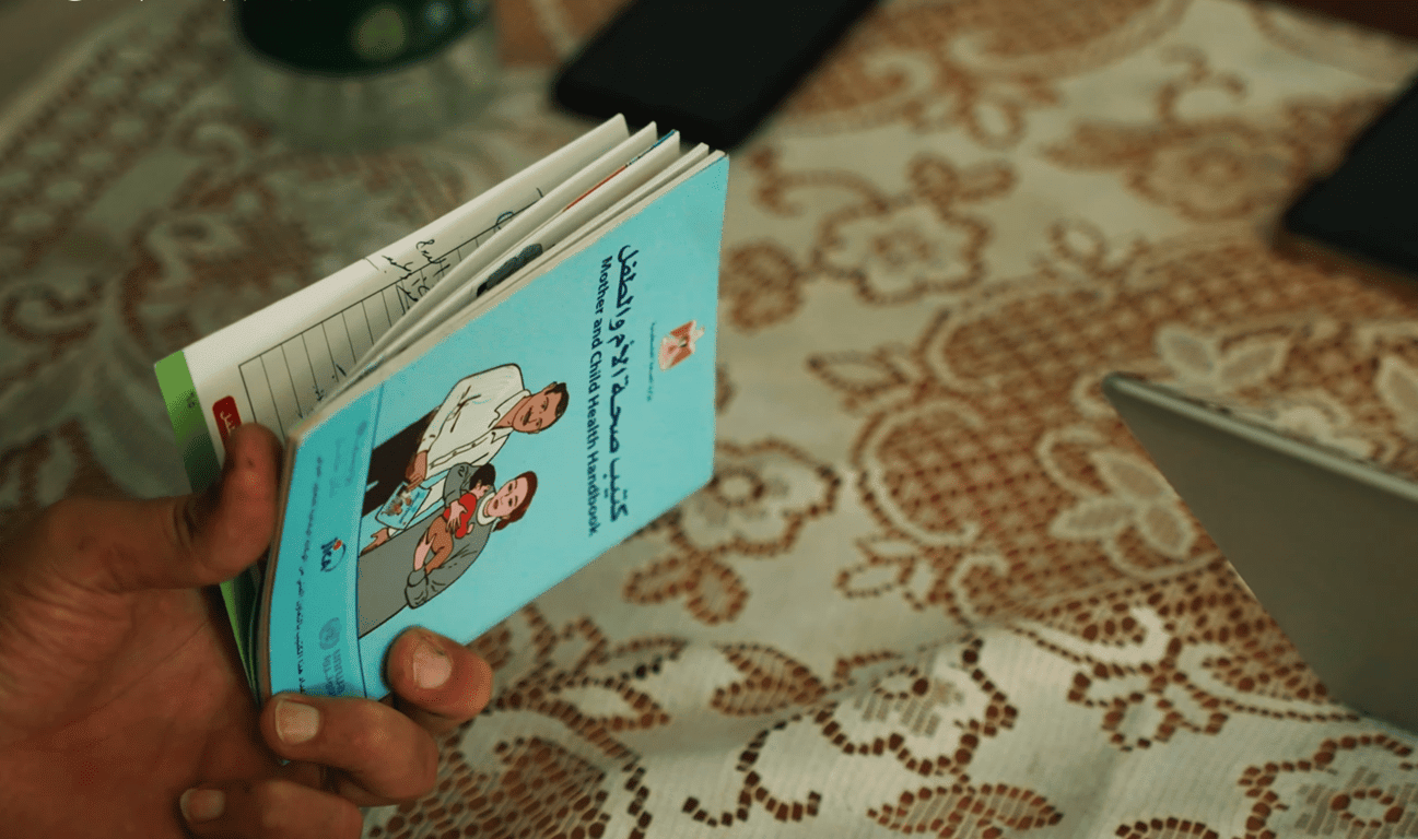 A hand holds an open Mother and Child Health Handbook in Arabic and English, depicting aid for Palestinian families via crypto donations, possibly using Solana.