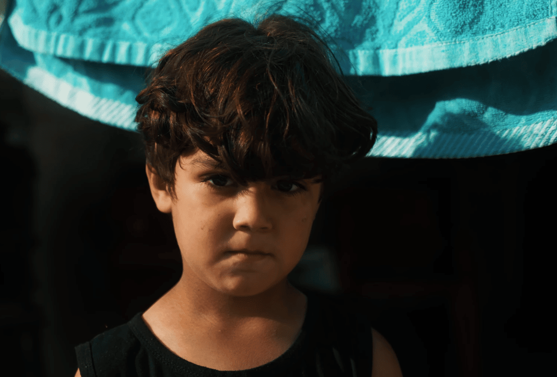 Close-up of a young Palestinian boy looking directly at the viewer, with a blue towel in the background. Your crypto Zakat donation, perhaps via BTC, can help save Palestinian children. Keywords: Zakat, Palestine, Muslims, crypto donation, children.