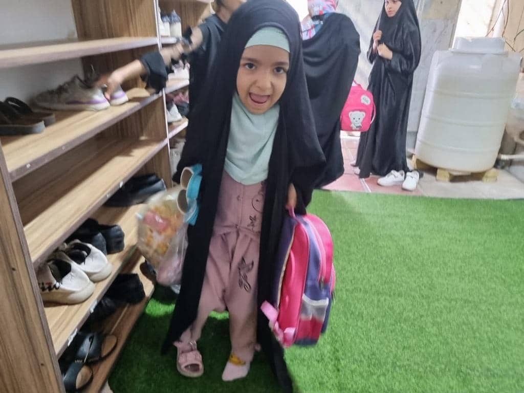 Young Palestinian girl with a backpack at a shoe rack smiling, representing crypto Zakat donations for aid in Palestine. TRX supported.