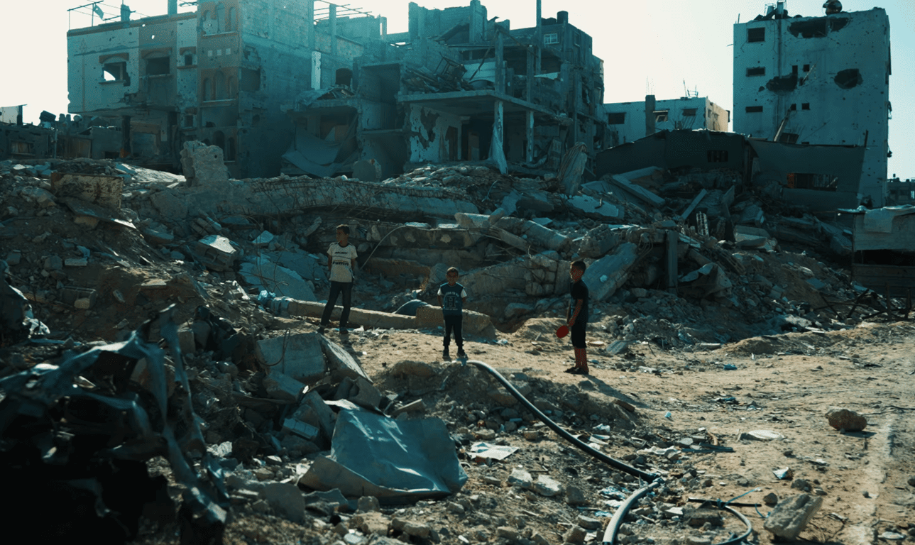 Three young boys stand amidst rubble and destroyed buildings, symbolizing the need for aid and relief efforts supported by BTC donations.