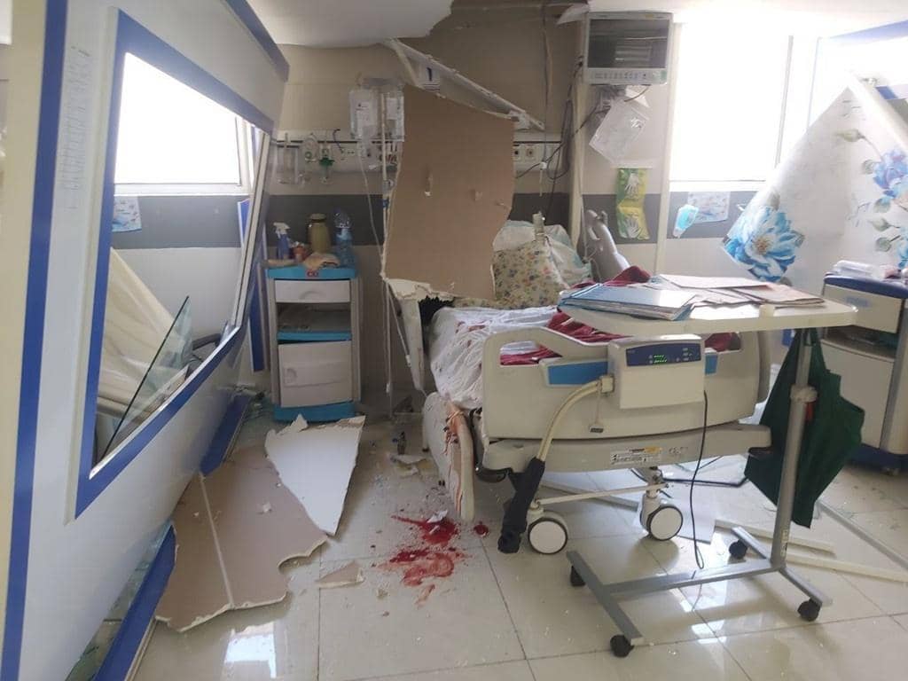 Damaged hospital room in Tehran due to war, showing shattered walls, an overturned medical cart, and a hospital bed splattered with blood. The destruction highlights urgent needs for aid, supported by fast cryptocurrency donations like Bitcoin.