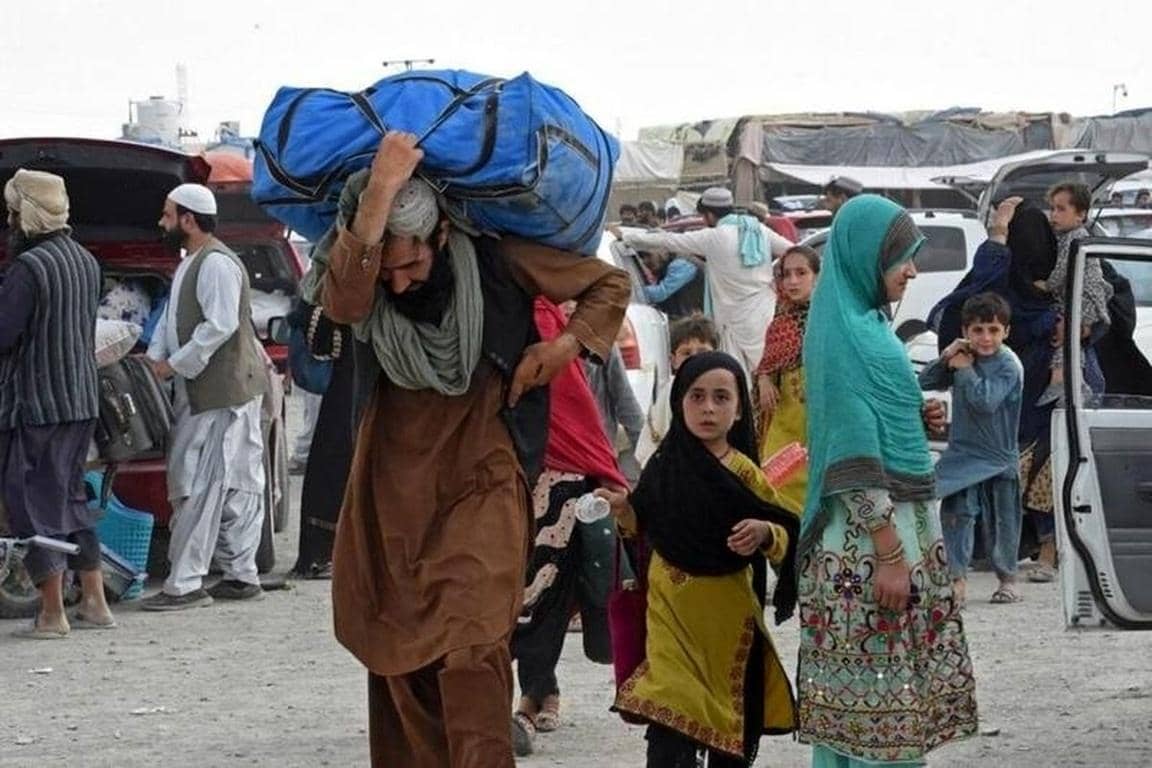 Man carrying heavy load, worried Afghan families at border, urgent aid via Solana, donations needed for food, water, shelter.