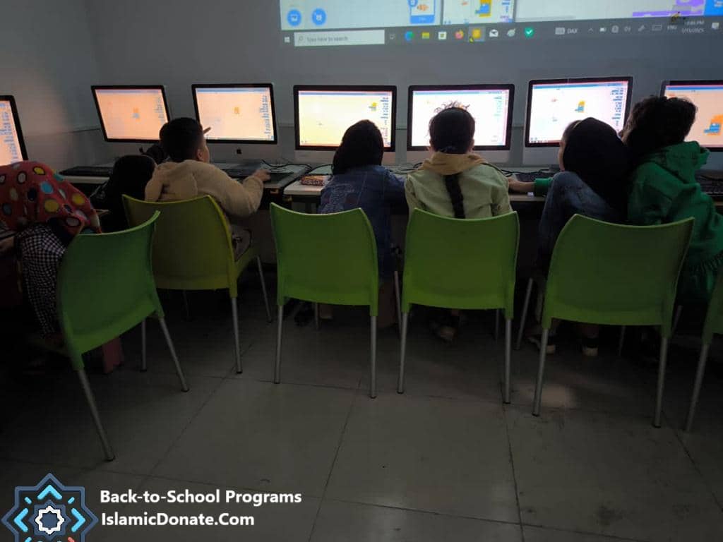 Children participating in an educational program, learning on computers, supported by anonymous crypto donations. A group of children are seated in front of multiple computer monitors, engaged in digital learning activities. This signifies a 'Back-to-School Program' initiative, enabling direct and secure donations via PayPal USD.