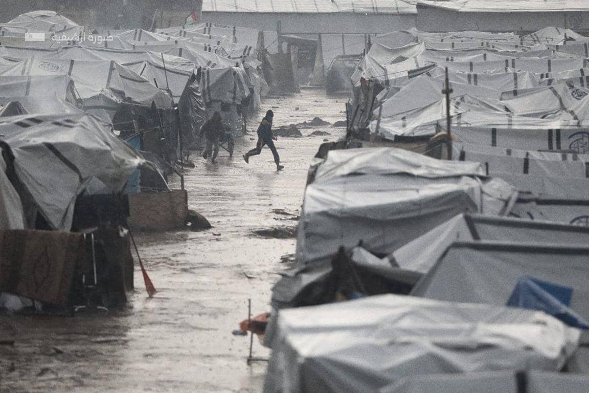 A child runs through a muddy, rain-soaked camp, seeking shelter amongst makeshift tents. This scene highlights the battle against cold and fear in Gaza, with aid supported by TRX donations.
