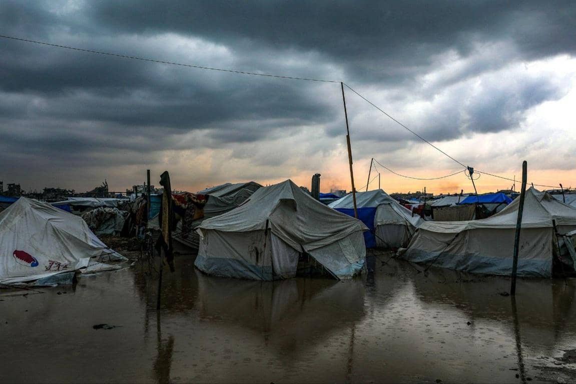 Flooded tent camp in Gaza under stormy skies, representing the urgent battle against winter crisis and the need for crypto donations from the Muslim Ummah for humanitarian aid.