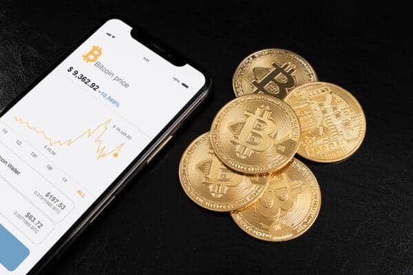 A smartphone displaying Bitcoin price chart next to several physical Bitcoin coins. This visual represents the growing ease of using cryptocurrency for transactions, with a focus on options like Bitcoin, Ethereum, and USDT for charitable aid.