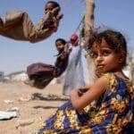 Child in a floral dress sits in a dusty, arid environment. Other children play on a makeshift swing in the background, suggesting relief efforts. Crypto donations are processed via BTC.