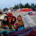 Children wrapped in blankets at a refugee camp in the Middle East, with tents in the background. Crypto donations support relief efforts.