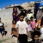 Children in a camp, some with vibrant clothing, others in worn attire. One child in the foreground smiles broadly.