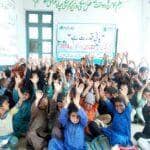 A group of children with their hands raised in a classroom setting, with a banner in the background reading "Muslim Aid". This scene depicts a charitable event or educational program, potentially funded by cryptocurrency donations. Keywords: Muslim Aid, children, education, charity, donation, crypto.
