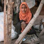 A young Yemeni girl in an orange hijab sits in a makeshift shelter, surrounded by sacks of World Food Program flour, reflecting the urgent need for humanitarian aid. Crypto donations via LTC offer a direct path to provide food security, health, and water, alleviating suffering in war-torn Yemen.