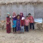 Children stand in front of a damaged shelter, one holding a box, implying humanitarian aid support via SOL crypto.