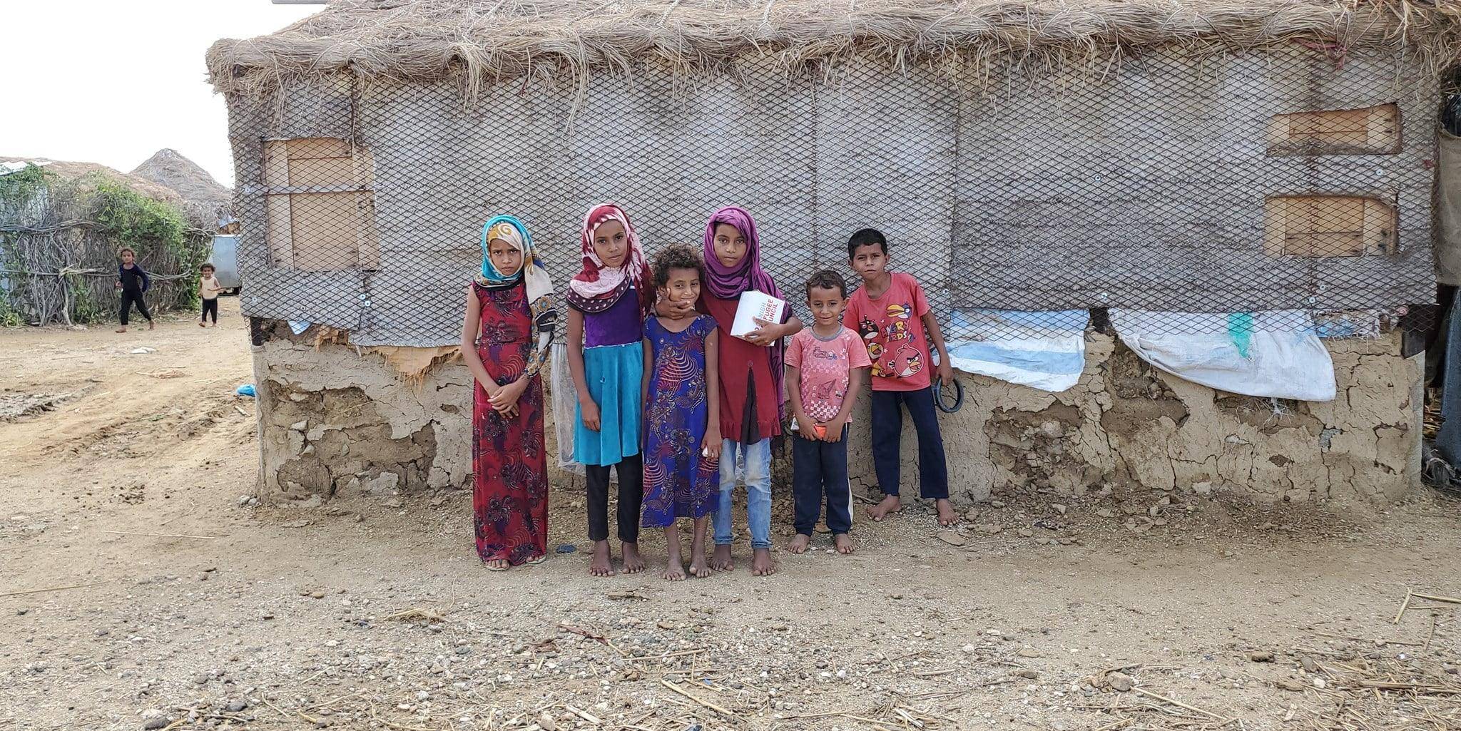 Children stand in front of a damaged shelter, one holding a box, implying humanitarian aid support via SOL crypto.