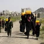 Children and women carrying water containers on their heads in a dusty village, with buildings in the background. Aid supported by ETH.