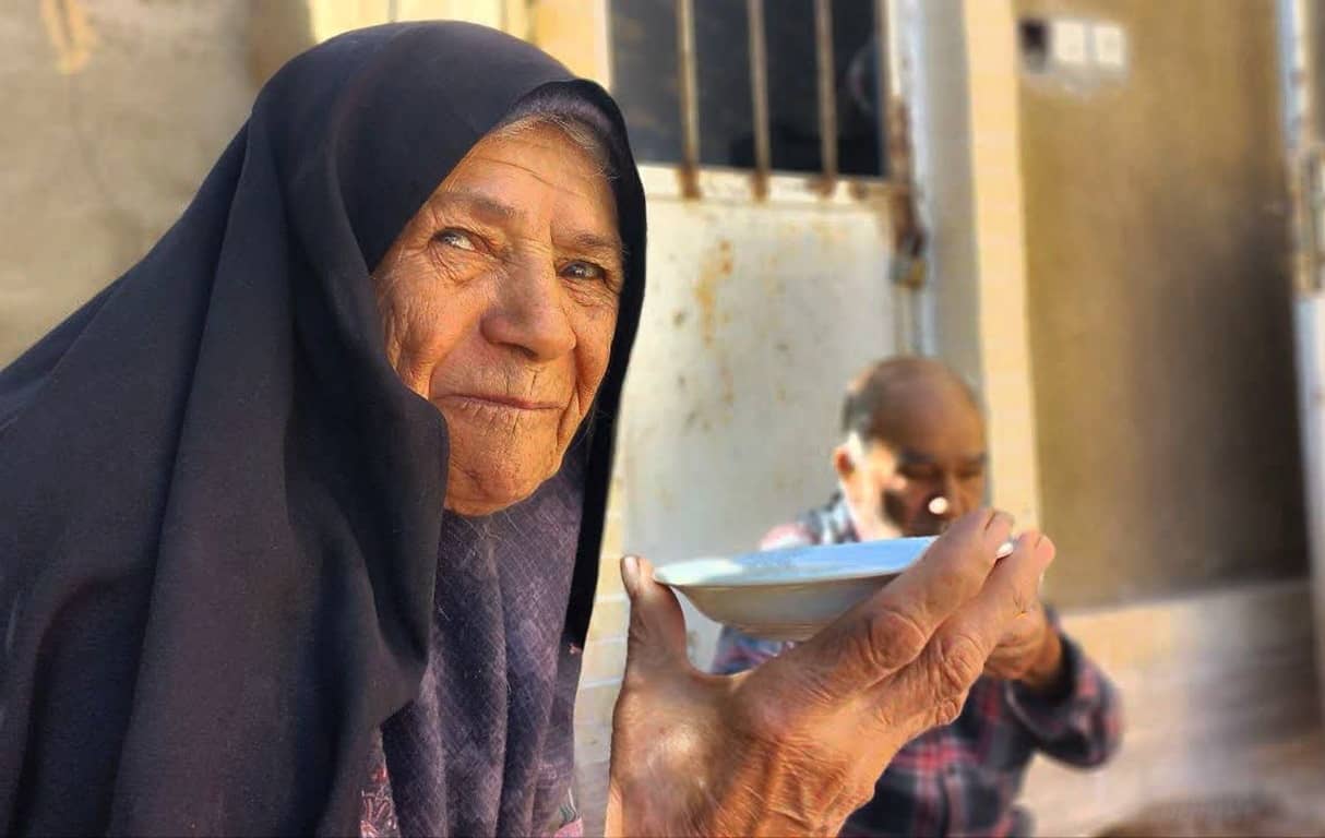 Elderly woman in hijab receiving a bowl of food, representing zakat donation to the poor and needy via cryptocurrency, with another person blurred in the background.