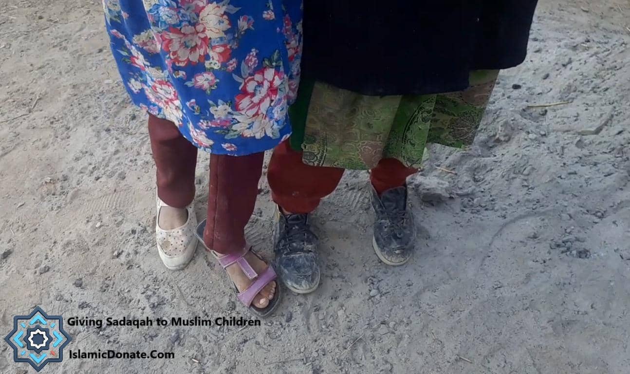 Close-up of children's feet wearing various shoes and sandals, standing on dusty ground. The image depicts the concept of giving Sadaqah, with a focus on providing aid and relief to Muslim children. Your donations, made via ETH, support essential needs like meals and family relief.