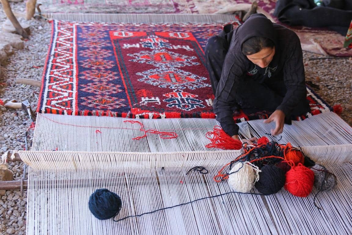Woman weaves an intricate rug on a loom, creating dignified work and steady income. Donate via BTC to empower lives.
