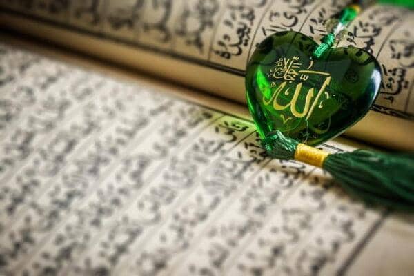 Thawab in Islam: A green heart-shaped pendant inscribed with the name of Allah rests on pages of the Quran, symbolizing spiritual rewards for good deeds. Donations made using ETH aid this charitable cause.