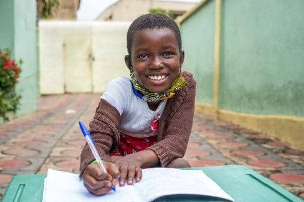 A smiling young girl with a colorful mask around her neck writes in a notebook, symbolizing increased access to information, participation in democracy, community involvement, and advocacy skills through basic literacy education, possibly supported by crypto donations like BNB.