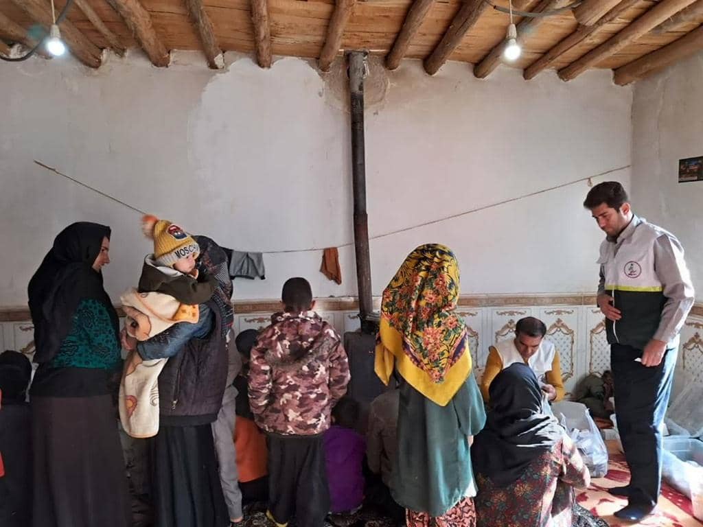 A man in a vest stands by a group of people, some children, in a modest room, distributing aid with the support of crypto donations via XRP.