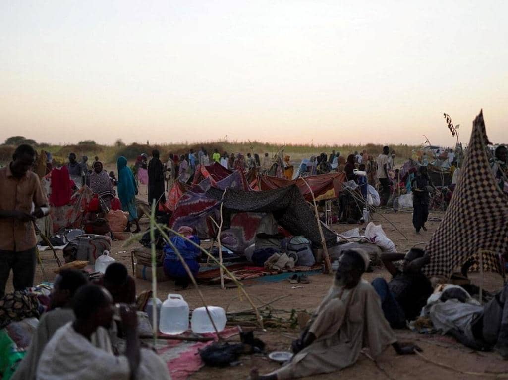 A crowded refugee camp with makeshift tents and people sitting on the ground, indicating urgent humanitarian aid needs in Sudan, supported by crypto donations like BTC.