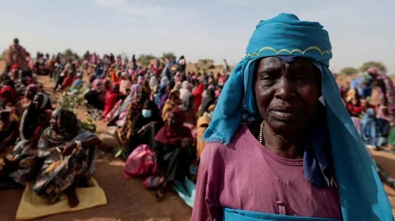 Displaced women and children gather in desert conditions, showcasing the urgent humanitarian crisis in Sudan and South Sudan, with a woman in foreground wearing a blue head covering. Donate Bitcoin to aid relief efforts.