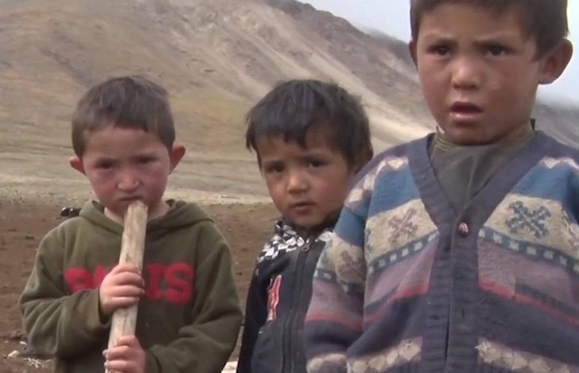 Three children looking at the camera in a desolate landscape, one holding a stick. Humanitarian aid for Afghanistan relief, including education and protection needs, can be donated via crypto like Ethereum.