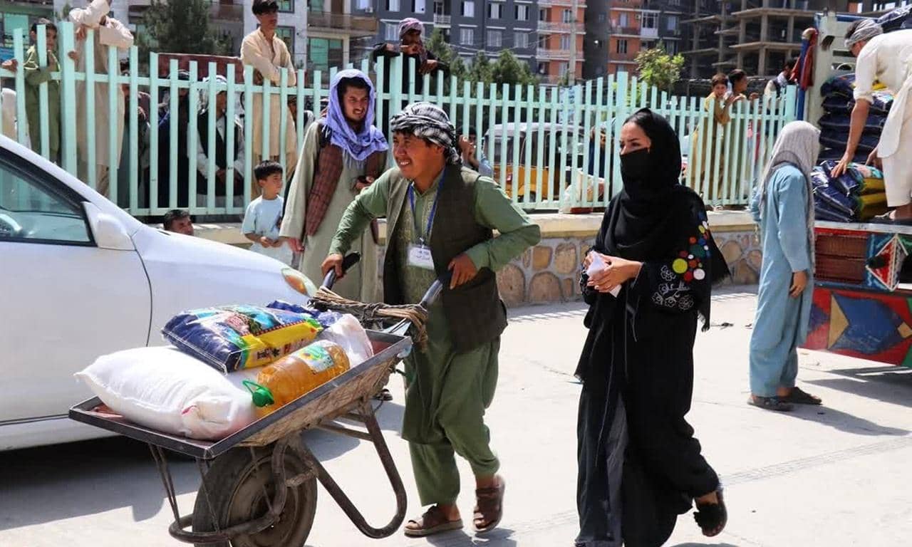 Afghan aid distribution showing a man pushing a wheelbarrow with food supplies, a woman in black hijab, and others receiving aid, supported by crypto donations via RLUSD.