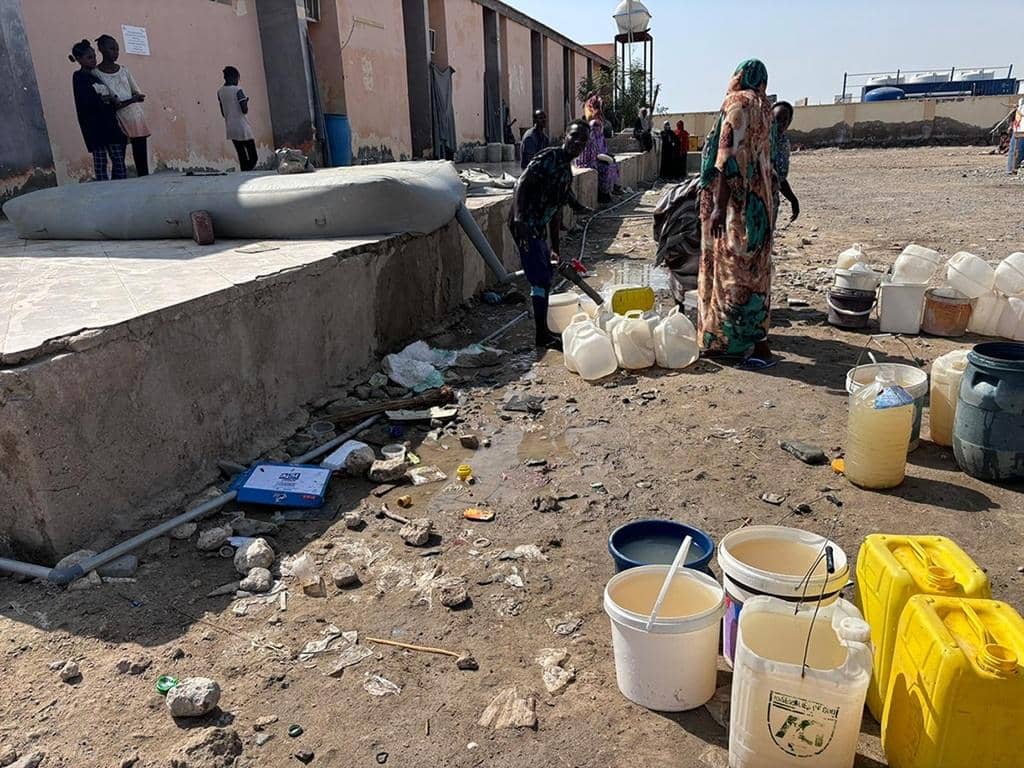People collecting water from a large tank in a crisis zone. This aid is facilitated by crypto donations, including BTC, offering essential relief like food and medical care.