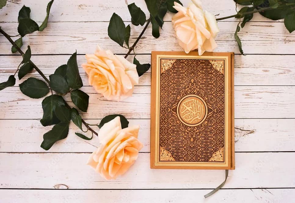 A decorative Quran is laid on a white wooden surface with several soft pink roses and green leaves, symbolizing a crypto-supported donation of the Holy Quran via USDC.