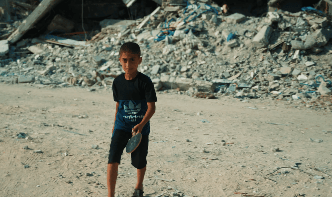 Orphan boy walks through rubble holding a ping pong paddle, symbolizing resilience and the need for support from crypto donations via RLUSD, ensuring food, shelter, and education.
