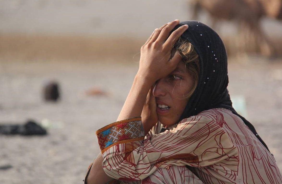 Woman with headscarf and patterned clothing, hands on forehead, appearing distressed. Aid via crypto, like Solana. Zakat al-Fitr, Ramadan charity, needy families, Islamic giving.